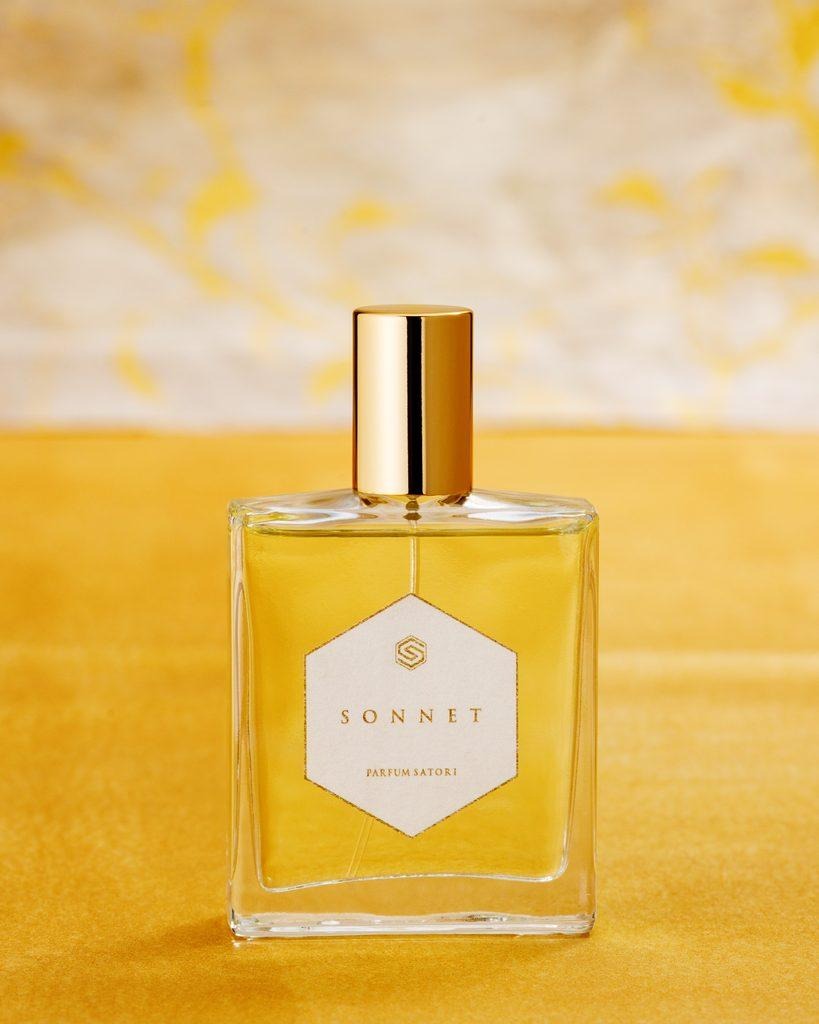 An elegant clear glass bottle of SONNET perfume with a gold cap resting gracefully on textured warm yellow fabric.
